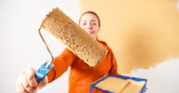 Personne tenant un rouleau et un bac de peinture. Elle est en train d'appliquer de la peinture anti-moisissure sur un mur blanc, qui devient beige.