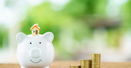 tirelire cochon surmontée d’une petite maison à côté de piles de pièces de monnaie pour illustrer une aide isolation combles proposée par Actimur.